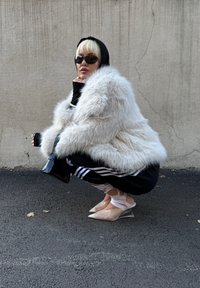 Bijela lažna krznena jakna, crni dugi rukavi majica, crne trenirke s bijelim trakama, prozirna bijela obuća, crne sunčane naočale, minimalistička crna torba.