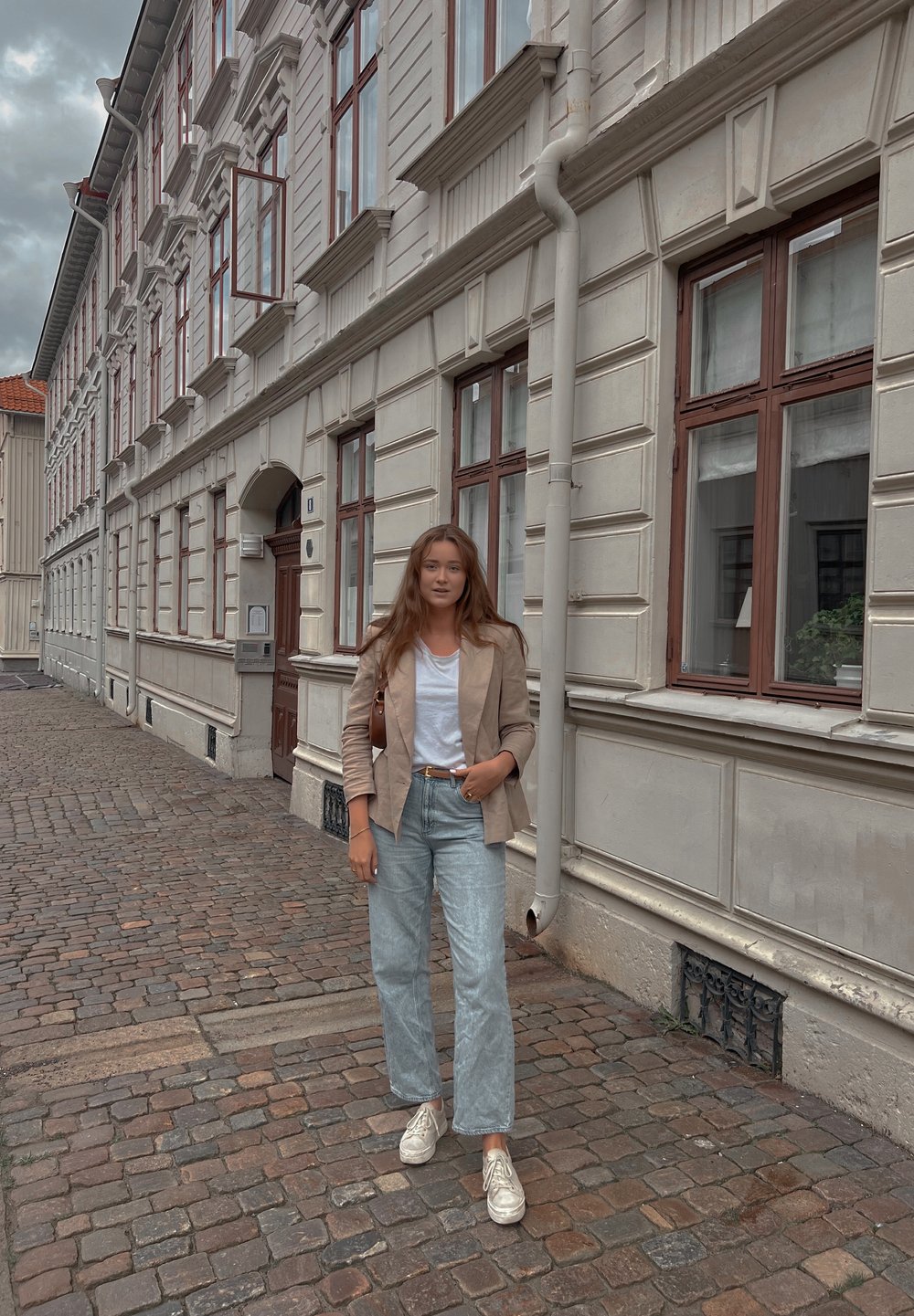 Απαλή μπεζ blazer πάνω από λευκό T-shirt, συνδυασμένο με ανοιχτό μπλε φαρδύ τζιν και λευκά sneakers; στέκεται σε πλακόστρωτο δρόμο με κτίρια.