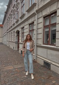 Απαλή μπεζ blazer πάνω από λευκό T-shirt, συνδυασμένο με ανοιχτό μπλε φαρδύ τζιν και λευκά sneakers; στέκεται σε πλακόστρωτο δρόμο με κτίρια.