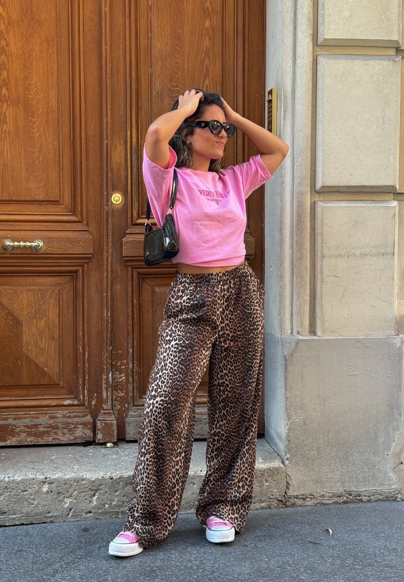 Ροδοκόκκινη κοντή μπλούζα με λογότυπο, καφέ παντελόνια σε λεοπάρ μοτίβο με φαρδιά γραμμή, μαύρη τσάντα χειρός και ροζ αθλητικά παπούτσια με λευκή σόλα μπροστά σε ξύλινη πόρτα.