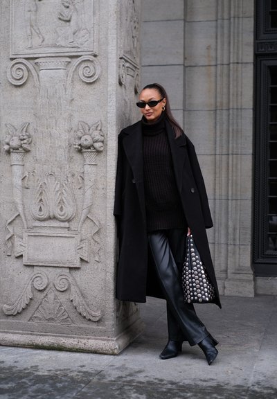 Abrigo negro sobre un suéter de cuello alto de punto, pantalones de cuero negros, botas de punta negra y un bolso con tachuelas contra un fondo de columna de piedra texturizada.