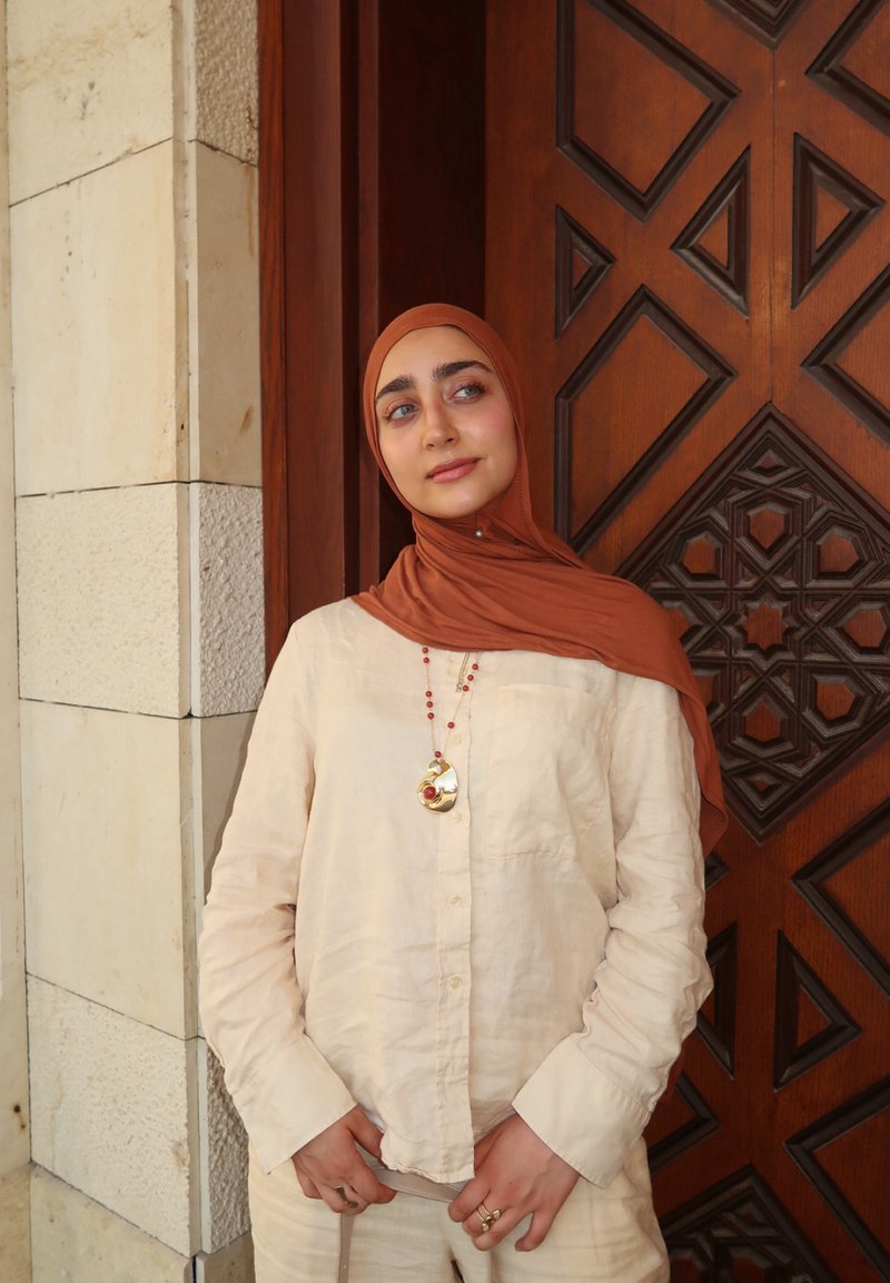 Junge Frau mit rostfarbenem Hijab und beigem Outfit steht neben einer verzierten Holztür und schaut leicht zur Seite mit einem ruhigen Ausdruck.