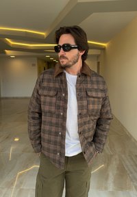 Brun ternet jakke med fløjlskrave, forsynet med trykknapper og to forlommer, båret over en hvid t-shirt og olivengr ønne cargobukser.