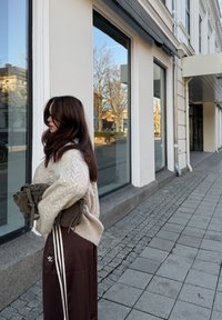 Lyse beige kabelstrikket sweater over brune atletikbukser med hvide striber. Bærer en mørkegrøn taske, står nær store vinduer på en fortov.