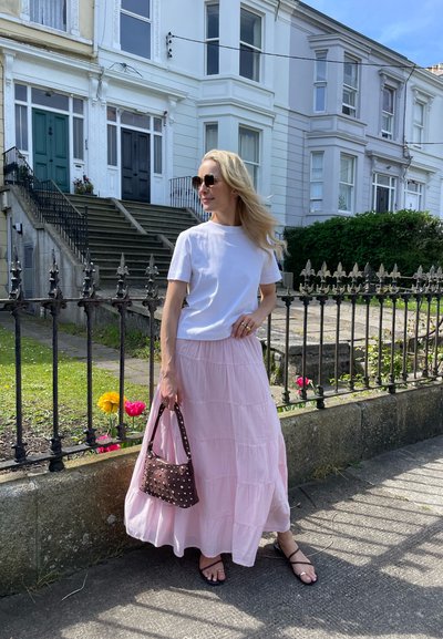 Camiseta blanca de manga corta, falda rosa con volantes, bolso marrón con tachuelas, sandalias negras. El fondo presenta un edificio blanco y una valla de hierro forjado.