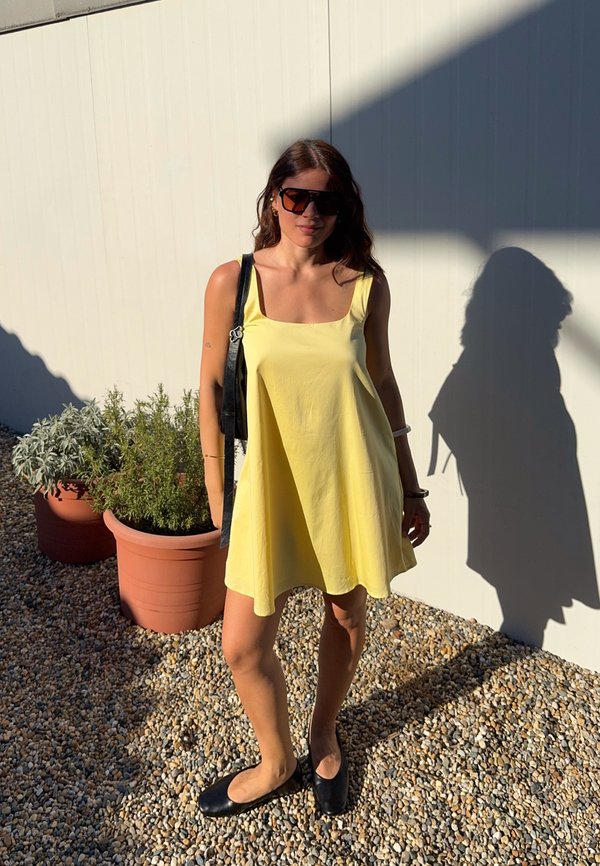 Femme portant une robe jaune, des chaussures noires et des lunettes de soleil, debout sur du gravier près de plantes en pot avec une ombre projetée sur un mur blanc derrière elle.