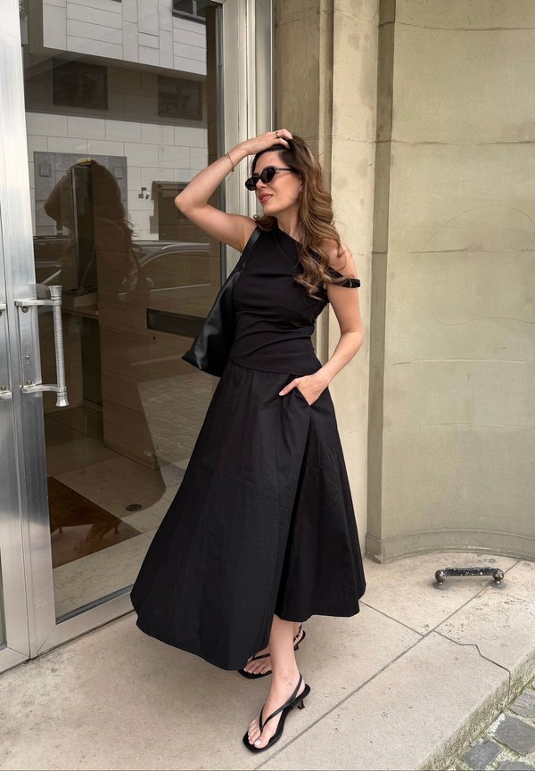 Femme aux longs cheveux portant des lunettes de soleil noires, une robe noire sans manches et des sandales noires, debout près d’une porte en verre, se touchant la tête d’une main.