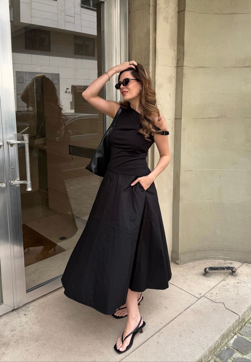 Frau mit langen Haaren, die eine schwarze Sonnenbrille, ein ärmelloses schwarzes Kleid und schwarze Sandalen trägt, steht nahe einer Glastür und berührt mit einer Hand ihren Kopf.