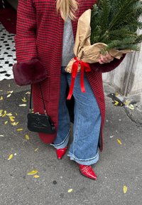 Manteau pied-de-poule rouge avec poignets en fausse fourrure bordeaux, pull gris, jean à jambes larges, talons rouges pointus cloutés, tenant un arbre en pot enveloppé de papier.