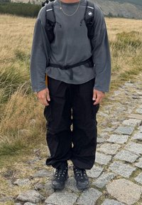 Osoba u crnim planinarskim čizmama, crnim cargo hlačama, sivom majicom dugih rukava i ruksakom, stoji na kaldrmisanoj stazi u travnatom krajoliku.