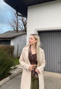 Šviesiai smėlinis ilgas paltas virš rudos trumpomis rankovėmis bluzonės, alyvuogių žalios kelnės ir maža ruda rankinė. Plaukai ilgūs ir tiesūs. Fone matomas pilkas garažo duris.