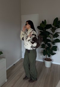 Brun og hvit flekket faux fur-jakke, grønne cargo-bukser, brune joggesko, med en brun veske, posert nær en potteplante innendørs.