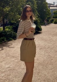 Pasiasta bluzka z krótkim rękawem w kolorze kremowym i brązowym, khaki szorty z wysokim stanem, trzymająca zielony napój w przezroczystym kubku. Naturalne otoczenie z zielenią.