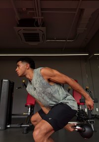 Person, der in einem Fitnessstudio eine Übung ausführt, trägt ein graues ärmelloses Shirt und schwarze Shorts, hält eine Kettlebell nahe der Fitnessgeräte.