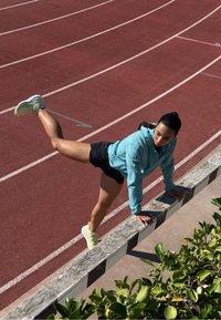 Athlet in einer hellblauen Zip-Jacke und schwarzen Shorts balanciert auf einer Holzbalustrade, hebt ein Bein nach oben, mit grünen Pflanzen in der Nähe.