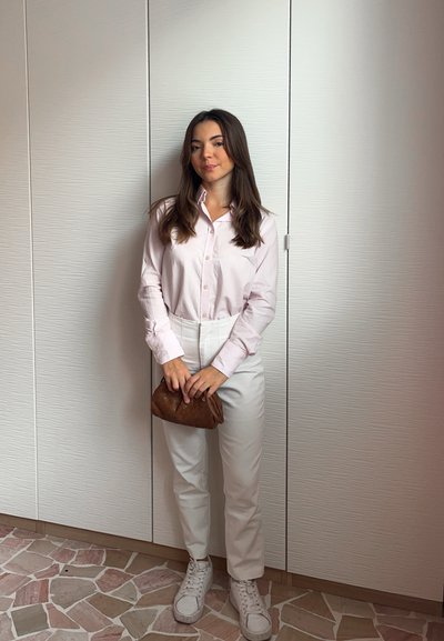 Camisa de manga larga rosa claro, pantalones blancos y bolso de mano trenzado marrón, combinados con zapatillas blancas. Texturas suaves y diseño ajustado.