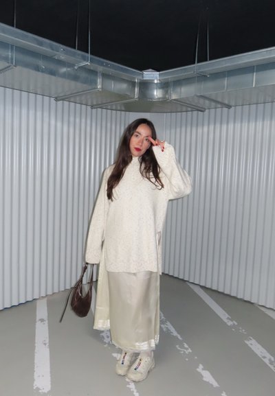 Mujer con un suéter grande color crema y falda, de pie en una esquina metálica e industrial, sosteniendo una bolsa marrón y haciendo el signo de la paz con uñas rojas.