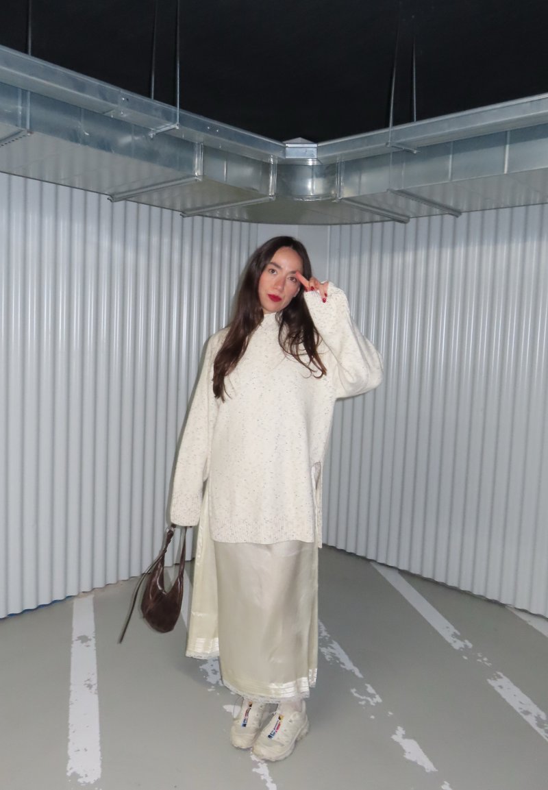 Frau in einem cremefarbenen, übergroßen Pullover und Rock steht in einer metallisch-industriellen Ecke, hält eine braune Tasche und zeigt mit roten Nägeln ein Peace-Zeichen.