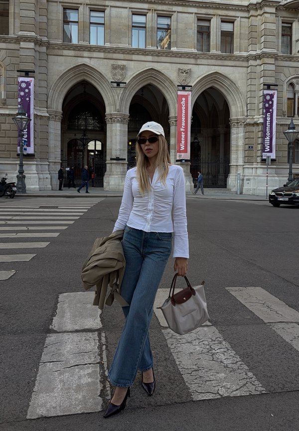 Kobieta w białej koszuli, czapce, okularach przeciwsłonecznych i dżinsach przechodzi przez przejście dla pieszych, trzymając beżową torbę i kurtkę przed budynkiem z arkadami.