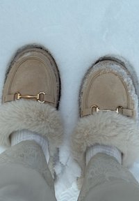 Mocassins en daim beige avec une bordure en fourrure douce, accents en métal doré, reposant sur un sol couvert de neige. Semelle texturée visible.