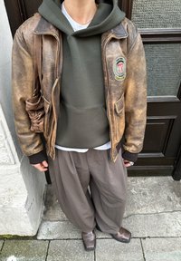 Barna bőrdzseki foltokkal, olívazöld kapucnis pulóver, fehér póló, bő szürke nadrág és barna, négyzet alakú orrú cipő, járdán állva.