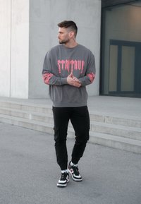 Sivá mikina s ružovým grafickým textom a vzorovanými rukávmi, skombinovaná s čiernymi cargo nohavicami a čierno-bielymi teniskami. Pozadie z betónu.