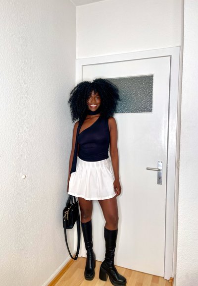 Top negro de un solo hombro, falda mini blanca plisada, botas de tacón negras hasta la rodilla y un bolso negro contra una pared y puerta blancas.