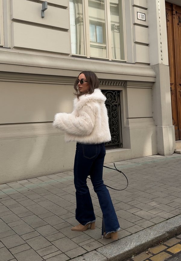 Giacca corta in pelliccia finta bianca, jeans a zampa d'elefante in denim scuro e stivaletti beige alla caviglia. L'outfit presenta un cappuccio e una vestibilità casual e rilassata.