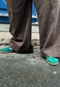Brune ternede bukser med brede ben og en glat tekstur; grønne ruskinds sneakers med hvide detaljer og en tekstureret sål.