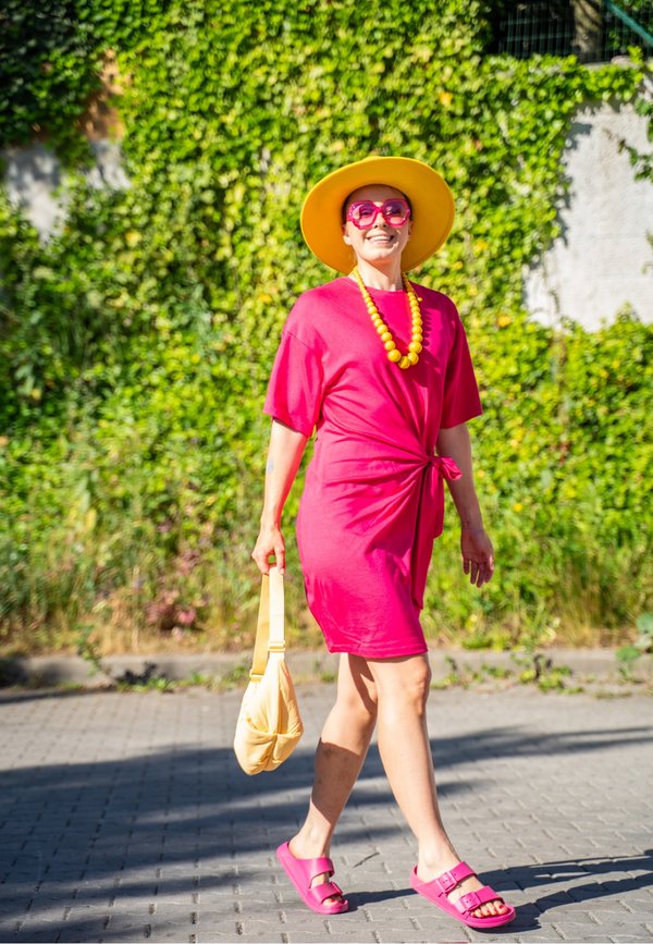 Frau in einem knallpinken Kleid, gelbem Hut und pinken Sandalen geht draußen mit einer gelben Tasche und einer Perlenkette, grüner blattiger Wand im Hintergrund.
