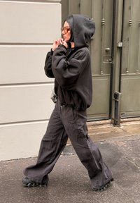 Oversized sort hættetrøje med hætte, kombineret med løstsiddende, mørkegrå cargobukser. Sorte sneakers med bobleformet sål; gennemsigtige solbriller.