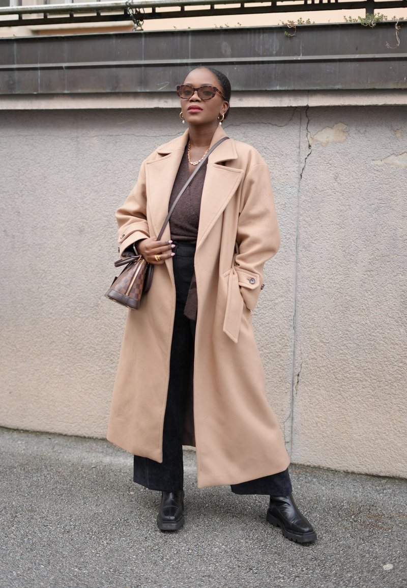 Μπεζ παλτό από μάλλινη μίξη με φαρδιά γιακά, κουμπιά, καφέ μπλούζα, μαύρα παντελόνια και χοντρές μαύρες μπότες. Καφέ τσάντα με σχέδιο.