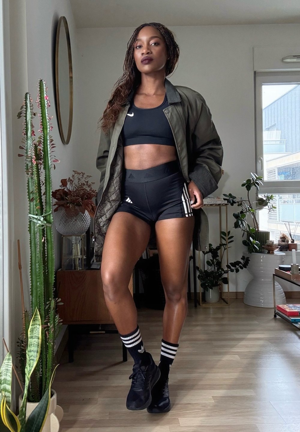 Femme portant un brassière de sport noire, un short Adidas noir, des chaussettes rayées et une veste verte ouverte, debout dans un salon moderne rempli de plantes.