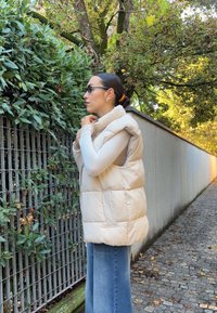 Šviesiai rudas pūkinis liemenė su aukštu apykakle, horizontaliu siuvinėjimo dizainu ir šoninėmis kišenėmis, vilkima virš šviesios ilgomis rankovėmis bluzonės, derinama su plačiomis džinsinėmis kelnėmis.