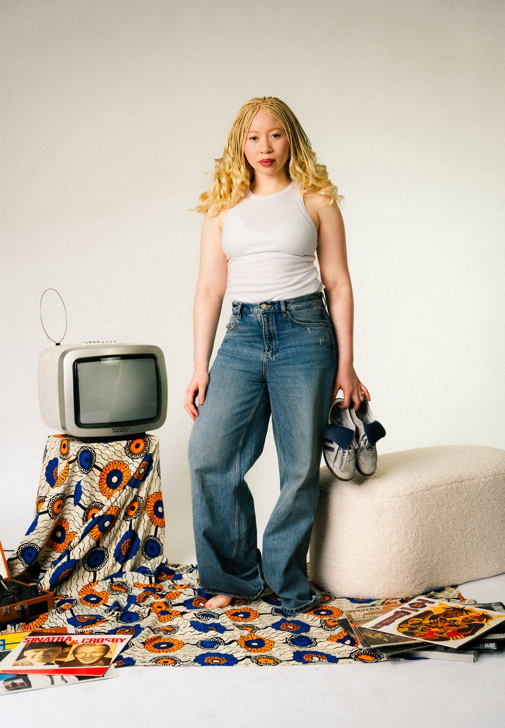 Vit tanktopp, blå lösa jeans, håller i grå sneakers. Bakgrunden visar en vintage TV på ett mönstrat tyg, omgiven av skivor och tidningar.