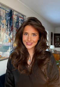 Jeune femme aux longs cheveux bruns ondulés souriant, portant un haut noir et un pendentif turquoise ; tableau abstrait et intérieur de maison en arrière-plan.