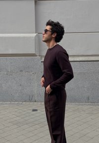 Model w ciemnorubinowym, fakturowanym swetrze i pasujących spodniach, stojący na tle jasnoszarej ściany o gładkiej powierzchni.