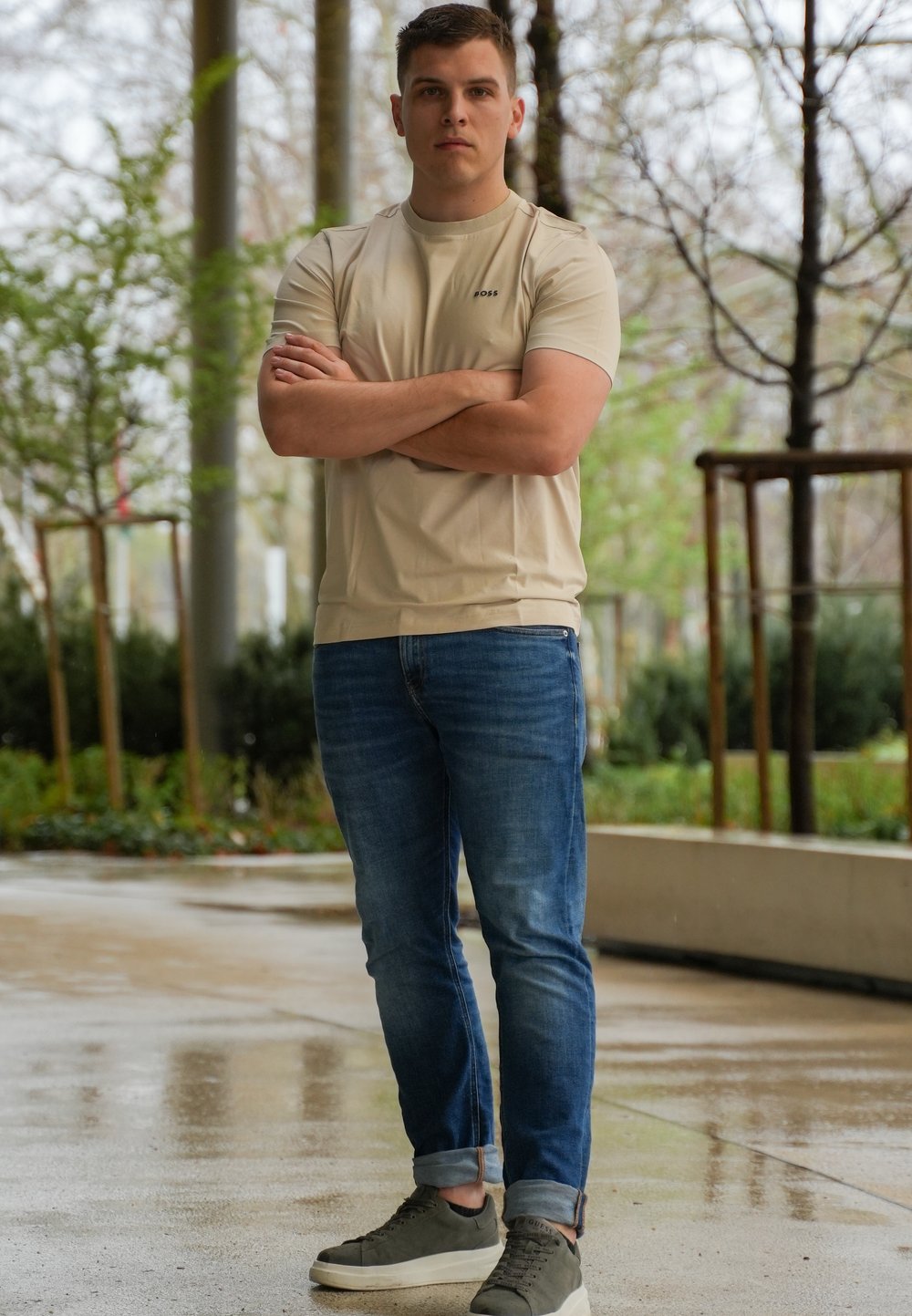 Beige tajta t-shirt med korta ärmar, blå denimjeans och gröna sneakers. Bakgrunden har grönska och blöt asfalt.