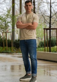 Beige tajta t-shirt med korta ärmar, blå denimjeans och gröna sneakers. Bakgrunden har grönska och blöt asfalt.