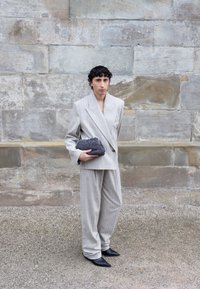 Persona con cabello rizado corto, vestida con un traje beige de gran tamaño y zapatos puntiagudos negros, sosteniendo un bolso de mano texturizado en gris oscuro.