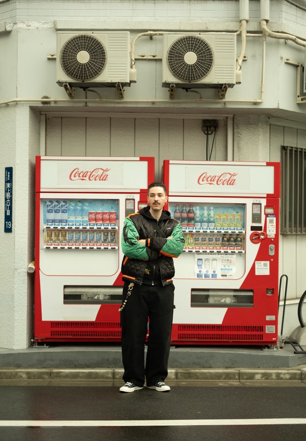 Mies seisoo käsivarret ristissä kahden Coca-Cola-automaatin edessä kaupunkikadulla ilmastointilaitteiden alla.