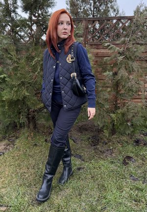 Joven con cabello rojo de pie sobre el césped cerca de arbustos y una cerca de madera, vistiendo un chaleco acolchado azul marino, pantalones y botas negras, llevando un pequeño bolso negro.