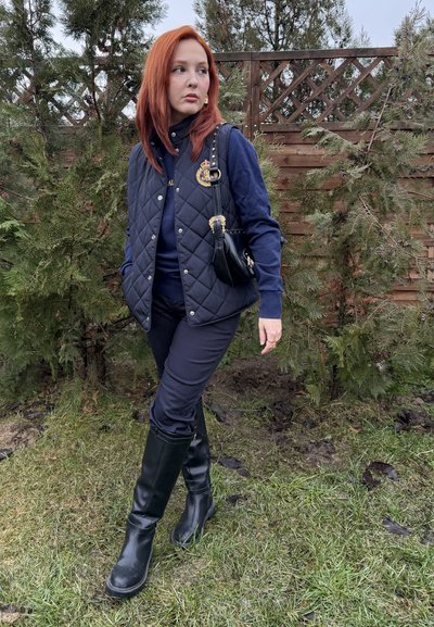 Joven con cabello rojo de pie sobre el césped cerca de arbustos y una cerca de madera, vistiendo un chaleco acolchado azul marino, pantalones y botas negras, llevando un pequeño bolso negro.