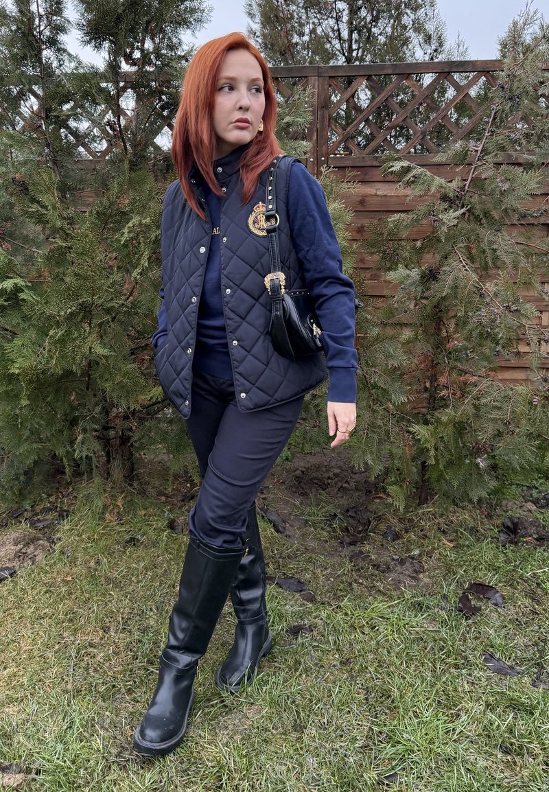 Jeune femme aux cheveux roux debout sur l'herbe près de buissons et d'une clôture en bois, portant une doudoune sans manches bleu marine, un pantalon et des bottes noires, avec un petit sac noir.