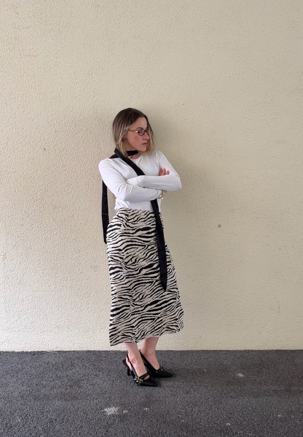 Kvinde med briller iført hvid langærmet trøje, sort tørklæde, zebra-mønstret nederdel og sorte hæle står med armene krydset mod en beige væg.