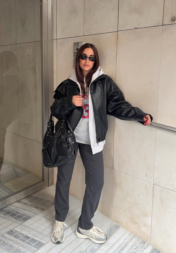 Femme portant une veste en cuir noire, un sweat à capuche gris, un pantalon foncé, des baskets blanches et des lunettes de soleil, portant un sac noir, debout à l'intérieur près d'un mur.