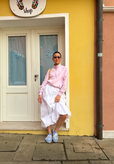 Chaqueta de cremallera rosa claro, falda blanca volumétrica y zapatillas azul claro. De pie cerca de una pared amarilla, con un cartel de tienda arriba y cortinas rayadas detrás.