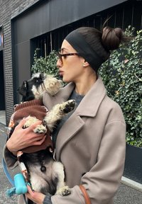 Femme en manteau beige et bandeau tenant un petit chien portant un pull marron, tous deux se regardant devant un mur couvert de lierre en plein air.