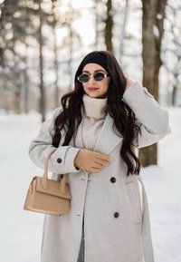 Femme aux longs cheveux foncés portant des lunettes de soleil, un bandeau noir, des gants beiges, un manteau et un pull, tenant un sac à main beige dans un décor extérieur enneigé.