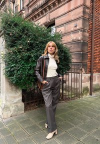 Braune Lederjacke, beiger Rollkragenpullover, graue Hose und hellbeige Schuhe. Grüner Busch-Hintergrund, Backstein- und Steingebäude.